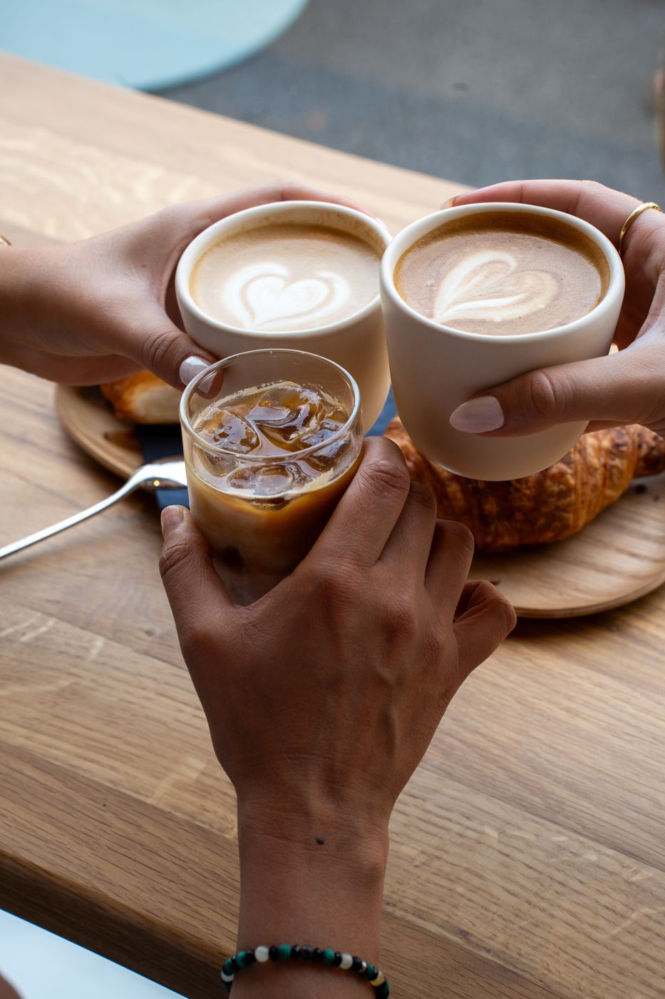 Drei Hände stossen mit Cappuccinos und einem Iced Latte über der Eichentheke an